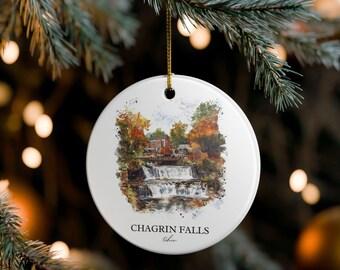 May include: A round white ceramic ornament featuring a watercolor-style illustration of Chagrin Falls, Ohio, with a waterfall, a bridge, and autumn-colored trees. The text CHAGRIN FALLS Ohio is printed in black at the bottom of the ornament.