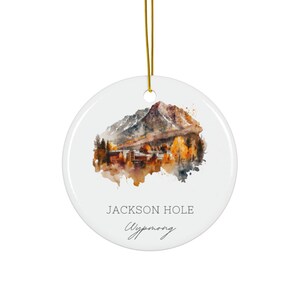 May include: A round, white ceramic ornament with a watercolor painting of a mountain landscape in autumn colors. The text "JACKSON HOLE Wyoming" is printed on the ornament, with a gold string for hanging.