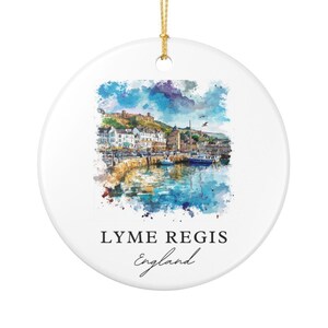 May include: A round white ceramic ornament featuring a watercolor illustration of the Lyme Regis harbor in England. The scene depicts buildings, boats, and water in blue, green, and brown tones. The text reads LYME REGIS England in black font.
