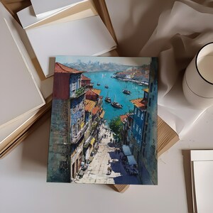 Puede incluir: Una pintura de acuarela de una ciudad costera europea. La obra de arte representa edificios con tejados de tejas rojas, un puerto azul con barcos y una calle con gente. La pintura se exhibe sobre una superficie blanca con otros materiales de arte.