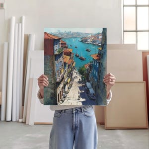 Puede incluir: Una pintura a la acuarela que representa un paisaje urbano europeo, con edificios con tejados naranjas y fachadas coloridas que conducen a un puerto con barcos. La pintura se sostiene frente a una persona.