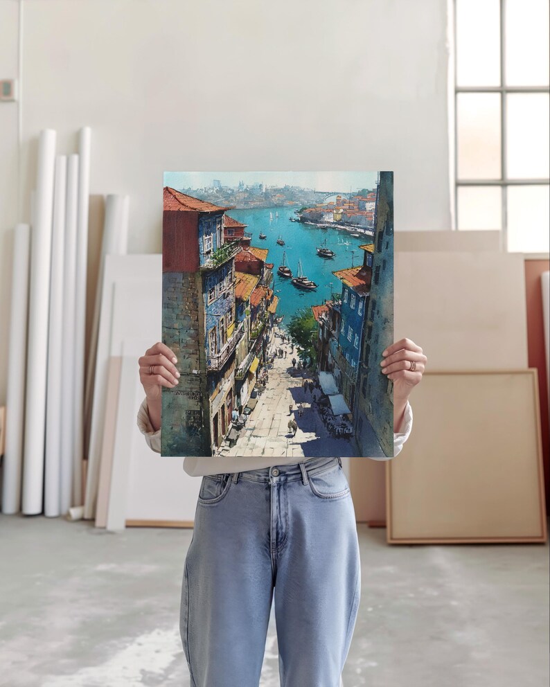 Puede incluir: Una pintura a la acuarela que representa un paisaje urbano europeo, con edificios con tejados naranjas y fachadas coloridas que conducen a un puerto con barcos. La pintura se sostiene frente a una persona.