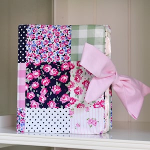 May include: A decorative book with a patchwork design featuring floral and polka dot patterns in pink, navy, and white. A large pink bow is tied on the side. The book is sitting on a white shelf.