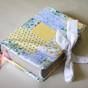 May include: A hardcover book with a patchwork fabric cover in yellow, blue, and white floral patterns. A white ribbon is tied around the book, and two colored ribbons serve as bookmarks. The book's edges are visible.