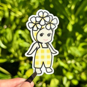 Puede incluir: Una pegatina con un personaje de dibujos animados con una corona de flores y un peto a cuadros amarillos. El personaje tiene una cara sonriente y est&aacute; delineado en negro. La pegatina est&aacute; sujeta con unas pinzas sobre un fondo verde borroso.