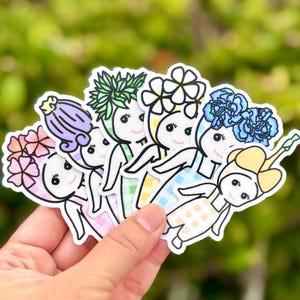 Puede incluir: Una colecci&oacute;n de pegatinas coloridas con personajes de dibujos animados con tocados florales. Cada pegatina tiene un dise&ntilde;o floral &uacute;nico en tonos rosa, morado, verde, amarillo y azul. Los personajes sonr&iacute;en y visten vestidos.