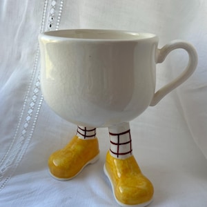 Könnte beinhalten: Ein weißer Keramikbecher mit einer glatten, abgerundeten Schale und einem einzelnen Henkel. Der Boden des Bechers ist als zwei Beine gestaltet, die weiße Socken mit einem dunkelroten Gittermuster und leuchtend gelbe Schuhe tragen.