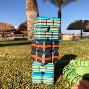 Könnte beinhalten: Ein Stapel aus drei bunten Fidget-Würfeln. Der obere Würfel ist blau, grün und pink mit Streifen. Der mittlere Würfel ist schwarz mit orangen Streifen. Der untere Würfel ist blau, grün und pink mit Streifen.