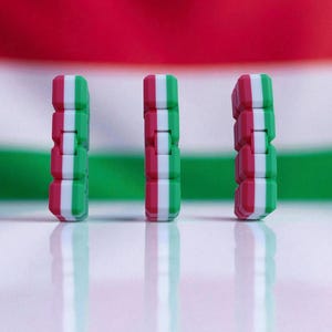 May include: Three vertical, stacked, rectangular objects with alternating red, white, and green segments, resembling a flag. The objects stand on a reflective white surface, with a blurred Hungarian flag in the background. The objects are a vibrant display of color.
