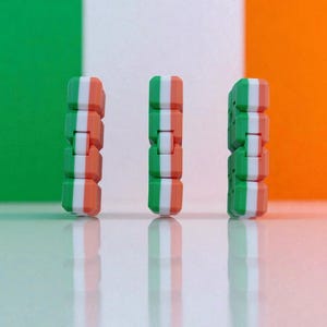 Op de afbeelding: Drie stapels kleine, rechthoekige blokken, elk met groene, witte en oranje strepen, staan tegen een achtergrond van de Ierse vlag. De blokken worden weerspiegeld op een glanzend oppervlak, waardoor een symmetrische compositie ontstaat.