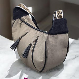 May include: A grey suede handbag with black trim and a leopard print strap. The bag has two tassels and a zip closure.