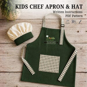 Può includere: Un set composto da grembiule da chef e cappello per bambini in verde scuro. Il grembiule presenta una tasca e lacci color crema con un motivo a triangoli verde scuro. Il testo ricamato recita Cookie Baker LUCAS. Il cappello ha la parte superiore crema.