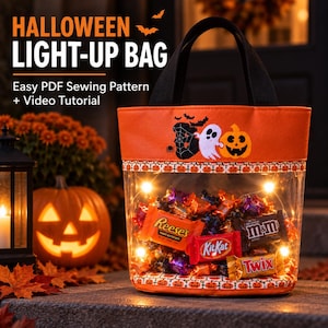 May include: An orange and clear Halloween light-up bag with black handles. The bag features a ghost, pumpkin, and spider web design with the text 'BOO'. It is filled with various candy bars and illuminated by internal string lights.