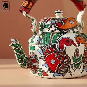 Könnte beinhalten: Ein weißer Teekessel aus Metall mit handbemalten, farbenfrohen Volkskunst-Illustrationen von Vögeln und Blumenmotiven in Rot, Grün, Orange und Blau. Der Kessel verfügt über einen passenden Deckel, einen geschwungenen Ausguss und einen roten Stoffgriff.