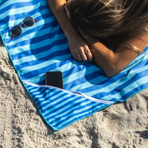 May include: A blue and white striped beach towel with a zippered pocket that holds a smartphone. The towel is lying on a sandy beach.