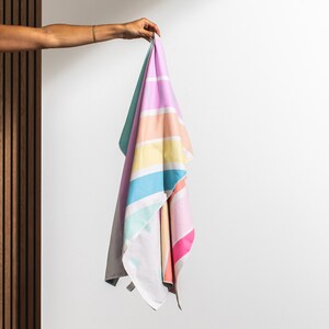 May include: A colorful striped beach towel with a rainbow pattern in shades of pink, blue, yellow, and green. The towel is folded and held by a hand.