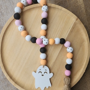 May include: A Halloween-themed door hanger with a white ghost cutout and a beaded garland. The garland is made of orange, pink, white, and black beads with white polka dots. The garland has a brown tassel.
