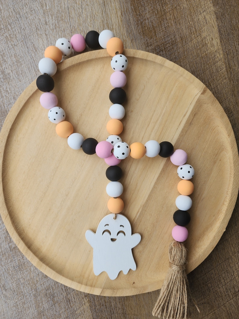 May include: A Halloween-themed door hanger with a white ghost cutout and a beaded garland. The garland is made of orange, pink, white, and black beads with white polka dots. The garland has a brown tassel.