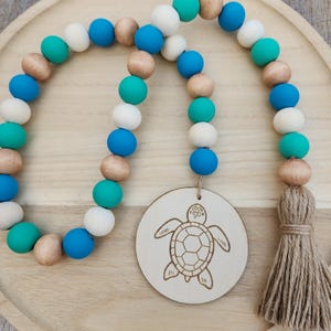 May include: A decorative wooden bead garland with a beach theme. The garland features teal, white, blue, and natural wood beads. A wooden disc with a laser-etched turtle design hangs from the garland, and a jute tassel is attached to the end.
