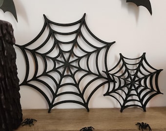 May include: Two black spider web cutouts with a radial design featuring interconnected concentric rings and spokes. The larger web is positioned to the left, while the smaller web is to the right. Both items are flat, matte black, and display a symmetrical, geometric pattern.