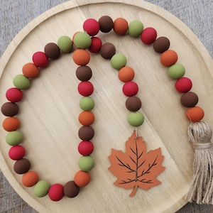 May include: A fall-themed wooden bead garland with red, brown, green, and orange beads. A wooden leaf charm hangs from the garland, and a natural fiber tassel is at the end. The garland is displayed on a wooden tray.