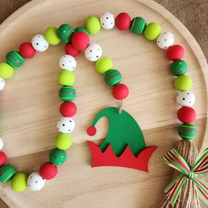 May include: A festive Christmas garland with red, green, and white wooden beads. The garland features an elf hat cutout and a jute tassel with a red and green striped bow. The beads are painted with stripes and polka dots.