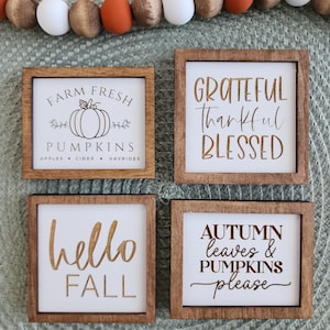 May include: Four square wooden signs with white backgrounds and dark brown frames. The signs feature black and gold text and graphics including Farm Fresh Pumpkins, Grateful Thankful Blessed, hello FALL, and AUTUMN leaves & PUMPKINS please.