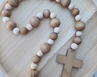 May include: A wooden beaded garland with a brown and white color scheme. The garland has a tassel at the end and is attached to a wooden cross.