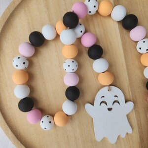 May include: A Halloween garland with black, white, pink, and orange wooden beads. The beads have black polka dots. A white ghost with a smiling face is hanging from the garland.