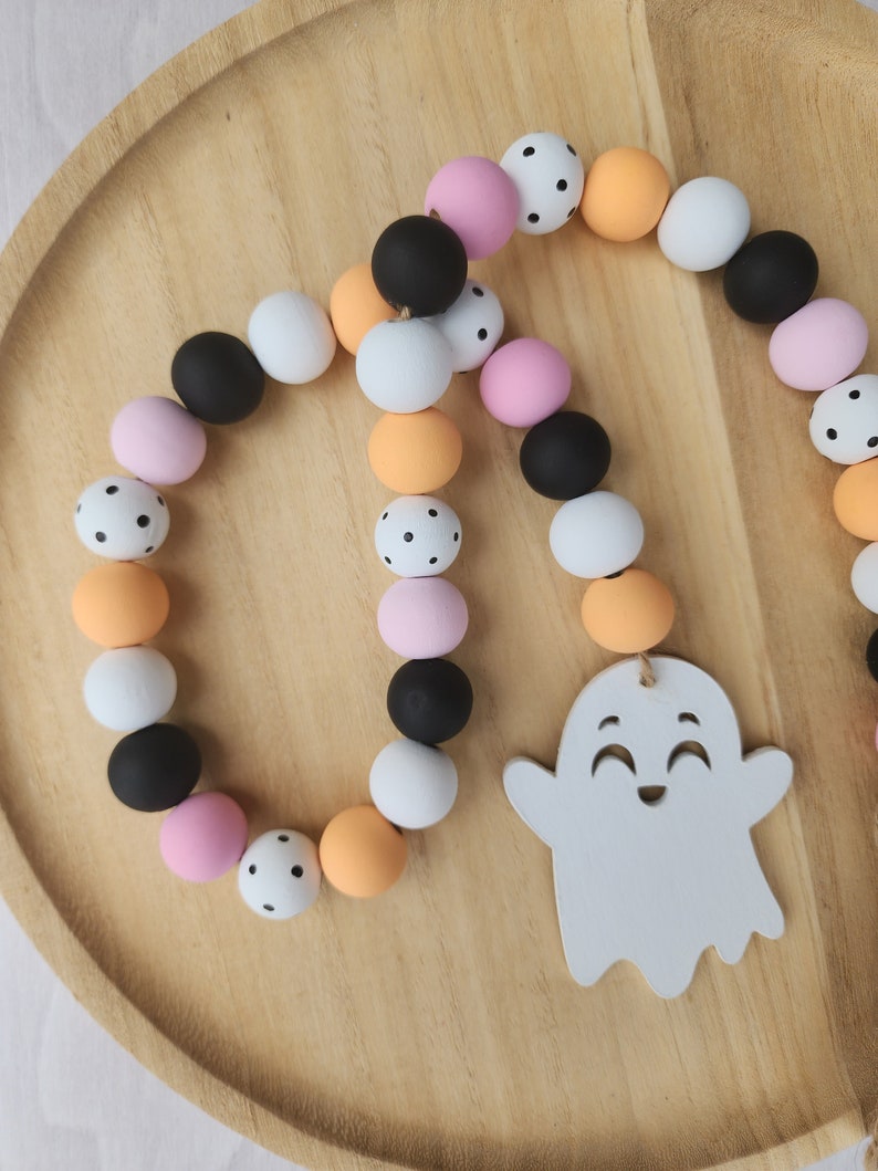May include: A Halloween garland with black, white, pink, and orange wooden beads. The beads have black polka dots. A white ghost with a smiling face is hanging from the garland.