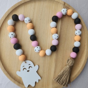 May include: A Halloween garland made of wooden beads in black, white, pink, and orange. The beads are decorated with polka dots. The garland has a wooden ghost charm with a smiling face and a brown tassel.