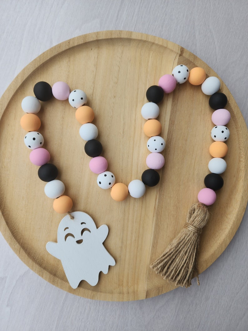 May include: A Halloween garland made of wooden beads in black, white, pink, and orange. The beads are decorated with polka dots. The garland has a wooden ghost charm with a smiling face and a brown tassel.