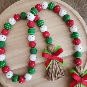 May include: A decorative garland featuring red, green, and white wooden beads. The beads are painted with polka dots and stripes. The garland includes jute tassels with red and green ribbon bows, perfect for holiday decor.