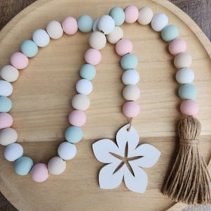 May include: A pastel-colored wooden bead garland with a white flower accent and a jute tassel. The beads are in shades of pink, blue, white, and beige. The garland is displayed on a wooden tray.