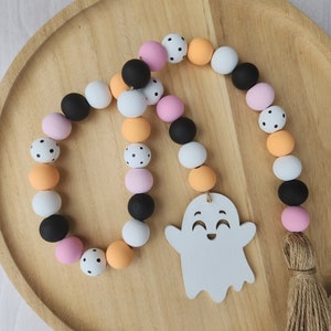 May include: A Halloween garland with a white ghost charm. The garland is made of wooden beads in black, white, orange, and pink with black polka dots. The garland has a brown tassel.