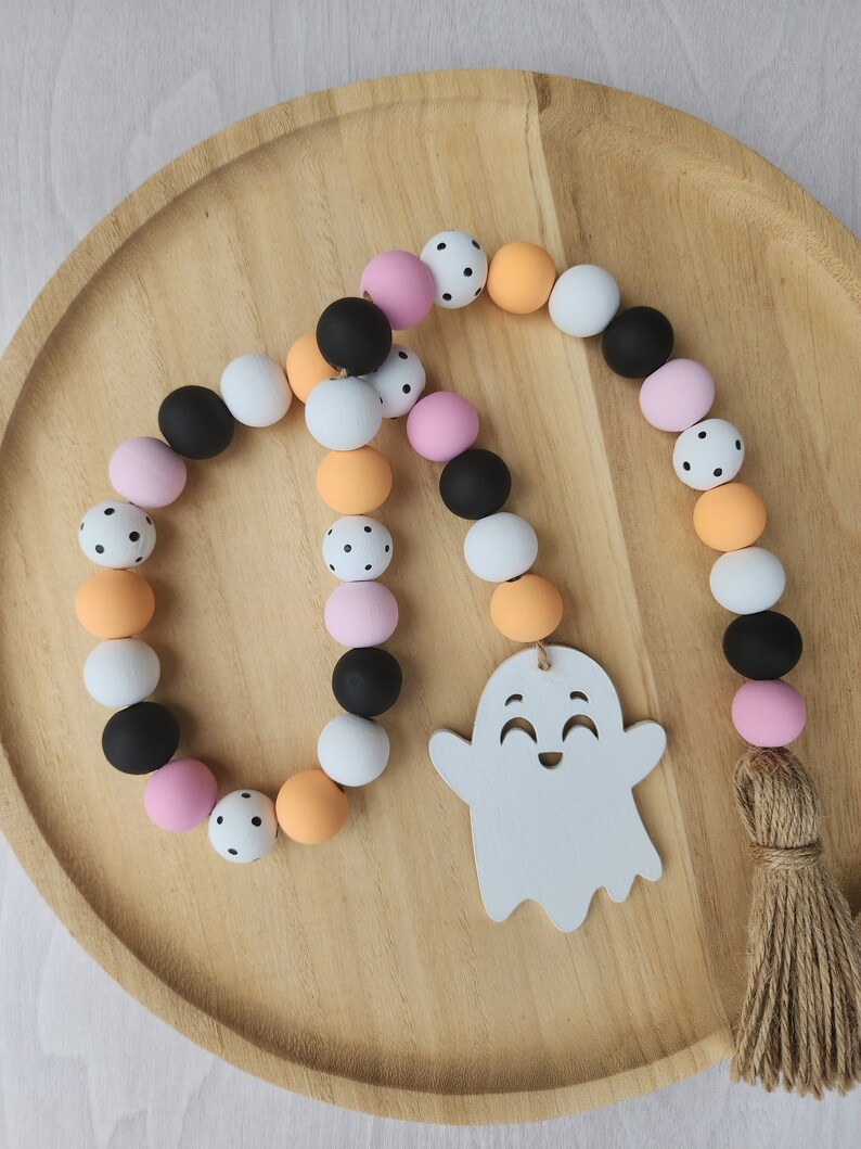 May include: A Halloween garland with a white ghost charm. The garland is made of wooden beads in black, white, orange, and pink with black polka dots. The garland has a brown tassel.