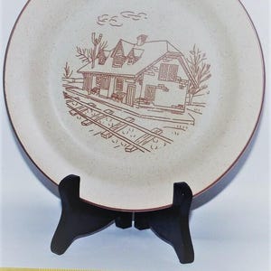May include: A round, speckled beige ceramic plate featuring a brown line-art illustration of a train station building and railroad tracks. A yellow measuring tape at the base shows the plate has a diameter of approximately 10.5 inches.