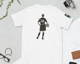 May include: White t-shirt with a black and white illustration of a soccer player on the back. The player is wearing a jersey with the number 23 on it and is holding a soccer ball.