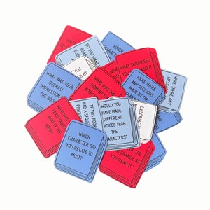 May include: A pile of red, white, and blue book-shaped cards, each printed with a different question about books and reading. Questions include "Which character did you relate to most?" and "Describe the message."