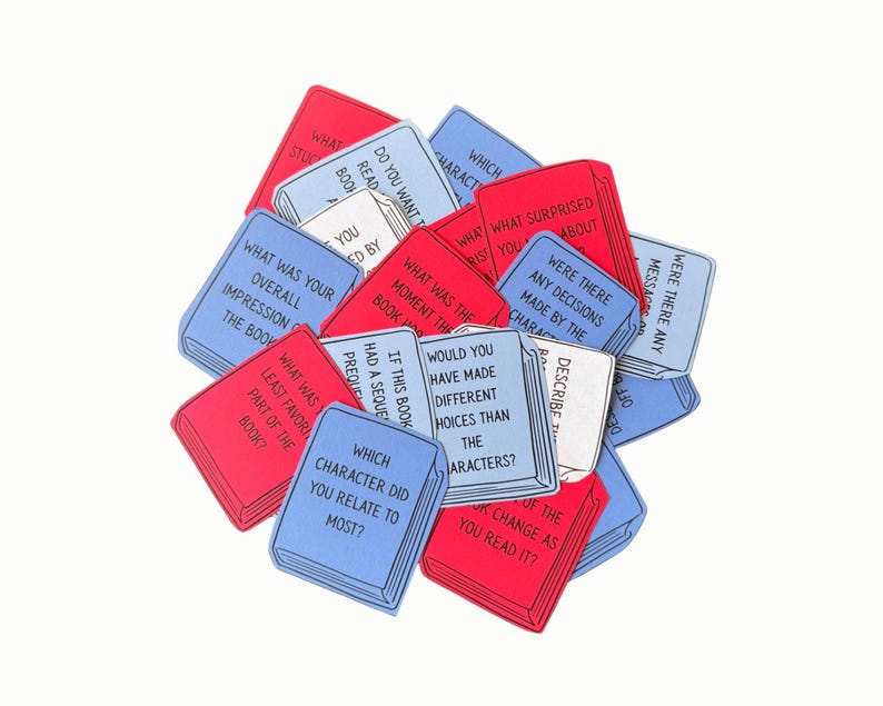 May include: A pile of red, white, and blue book-shaped cards, each printed with a different question about books and reading. Questions include "Which character did you relate to most?" and "Describe the message."