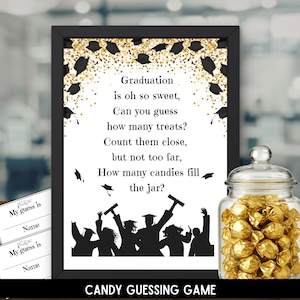 May include: A framed graduation-themed candy guessing game with a black frame, gold confetti, and a silhouette of graduates. The text reads "Graduation is oh so sweet... How many candies fill the jar?" A glass jar filled with gold-wrapped candies is next to the frame.