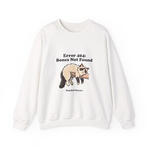May include: A white long-sleeve sweatshirt featuring a graphic of a cream and brown cat draped over an arm. The text above the cat reads "Error 404: Bones Not Found" and the text below the cat reads "Ragdoll Physics" in black lettering.