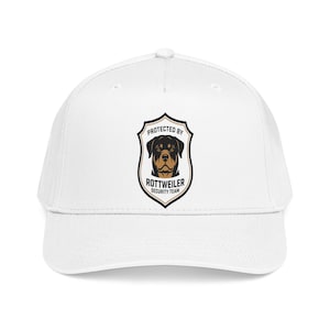 May include: A white baseball cap featuring a central shield-shaped graphic. The design includes a Rottweiler dog illustration with the text "PROTECTED BY ROTTWEILER SECURITY TEAM" printed in black and tan colors within the shield border.