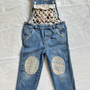 Puede incluir: Pantalón vaquero azul con peto, detalle de crochet blanco en el pecho y parches de tela a cuadros en las rodillas.