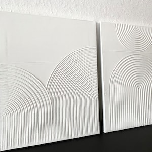 May include: Two white canvas wall art pieces with raised, textured, concentric circles in a geometric pattern.