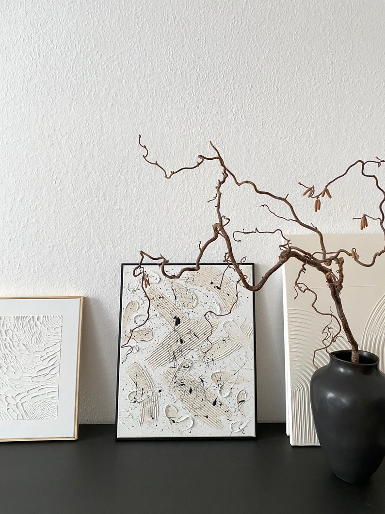 以下が含まれることがあります： 白い背景に白と黒の塗料で描かれた抽象画が3つ額装されています。絵画の前に、枯れ枝が入った花瓶があります。