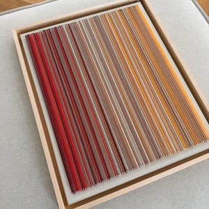 Könnte beinhalten: Ein gerahmtes Kunstwerk mit einem Farbverlauf aus roten, braunen und orangenen Fäden auf weißem Hintergrund. Die Fäden sind parallel angeordnet und erzeugen ein strukturiertes, abstraktes Design. Das Kunstwerk ist in einem hellen Holzrahmen gefasst.