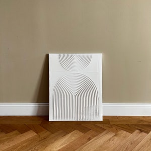 May include: White abstract wall art with a raised design of overlapping circles and arches. The art is on a beige wall with a wood floor.