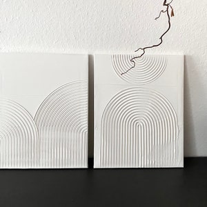 May include: Two white square canvases with raised, embossed, concentric circles and semi-circles in a geometric pattern.