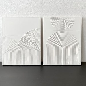 May include: Two white square canvases with raised, textured, white geometric shapes. The shapes are semi-circles and arcs, creating a modern abstract design.